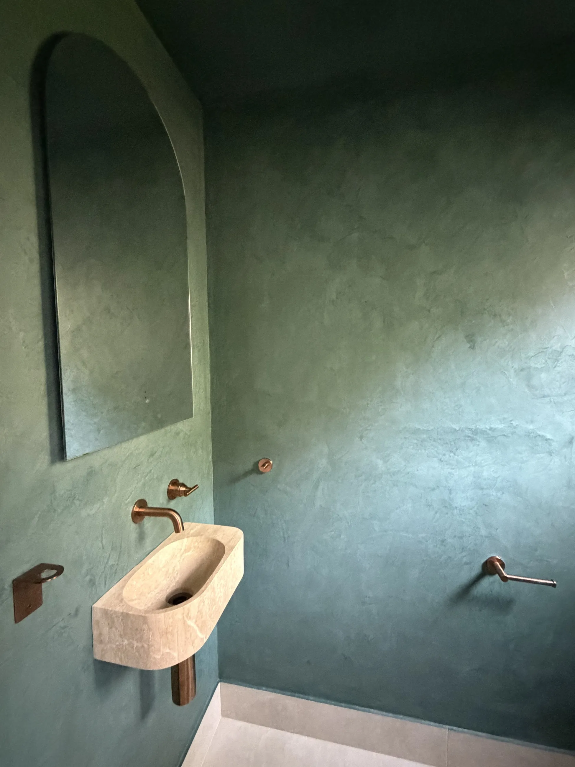 A powder room wall in a hand-applied decorative finish with soft, mottled depth in deep green, beside an arched mirror, a pale stone basin and brass tapware.